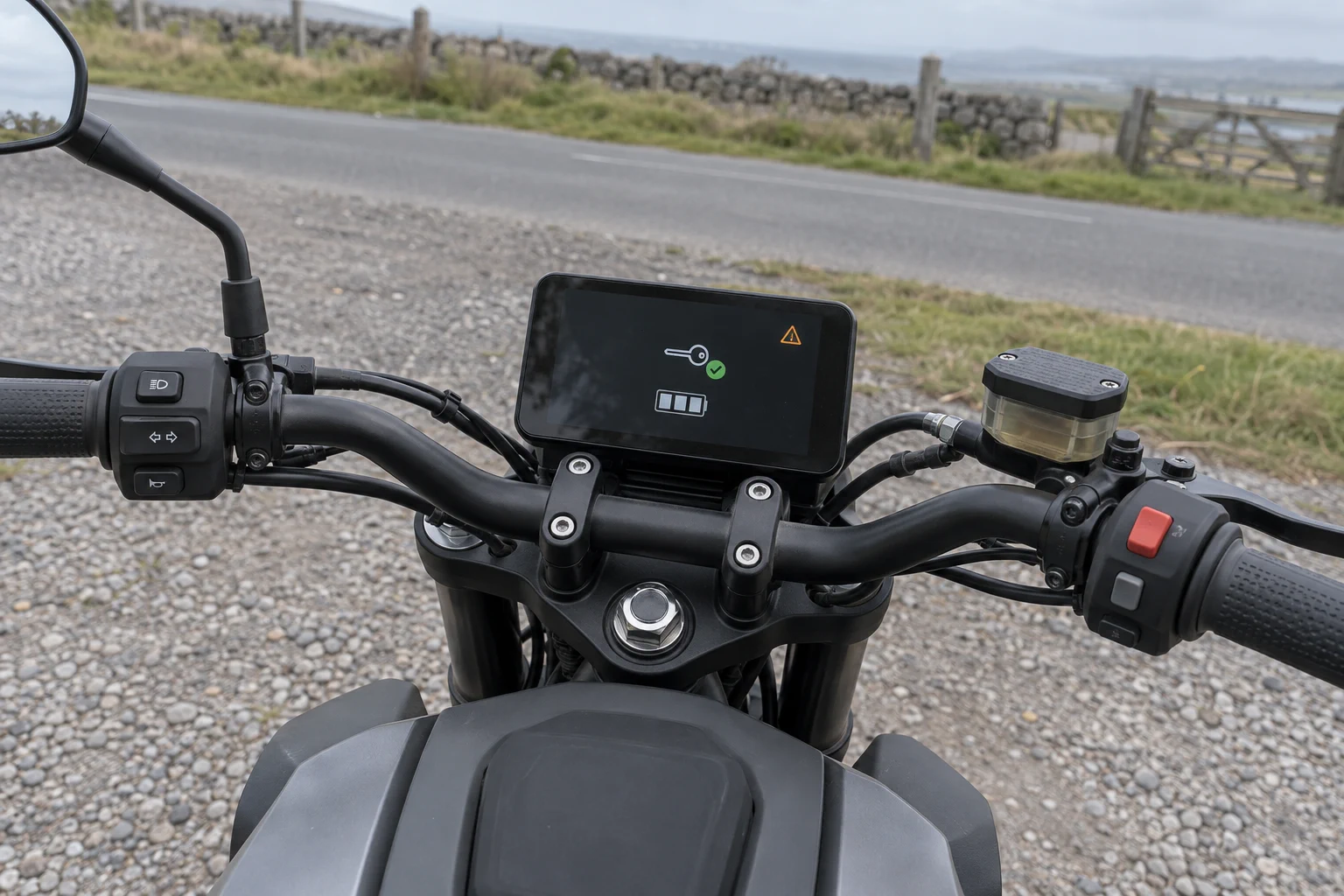 Parked motorcycle cockpit in overcast daylight, with a simple key recognition icon, battery segments and amber warning on its instrument display.